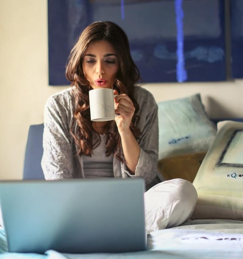 Brunette woman sits on bed with laptop and coffee
